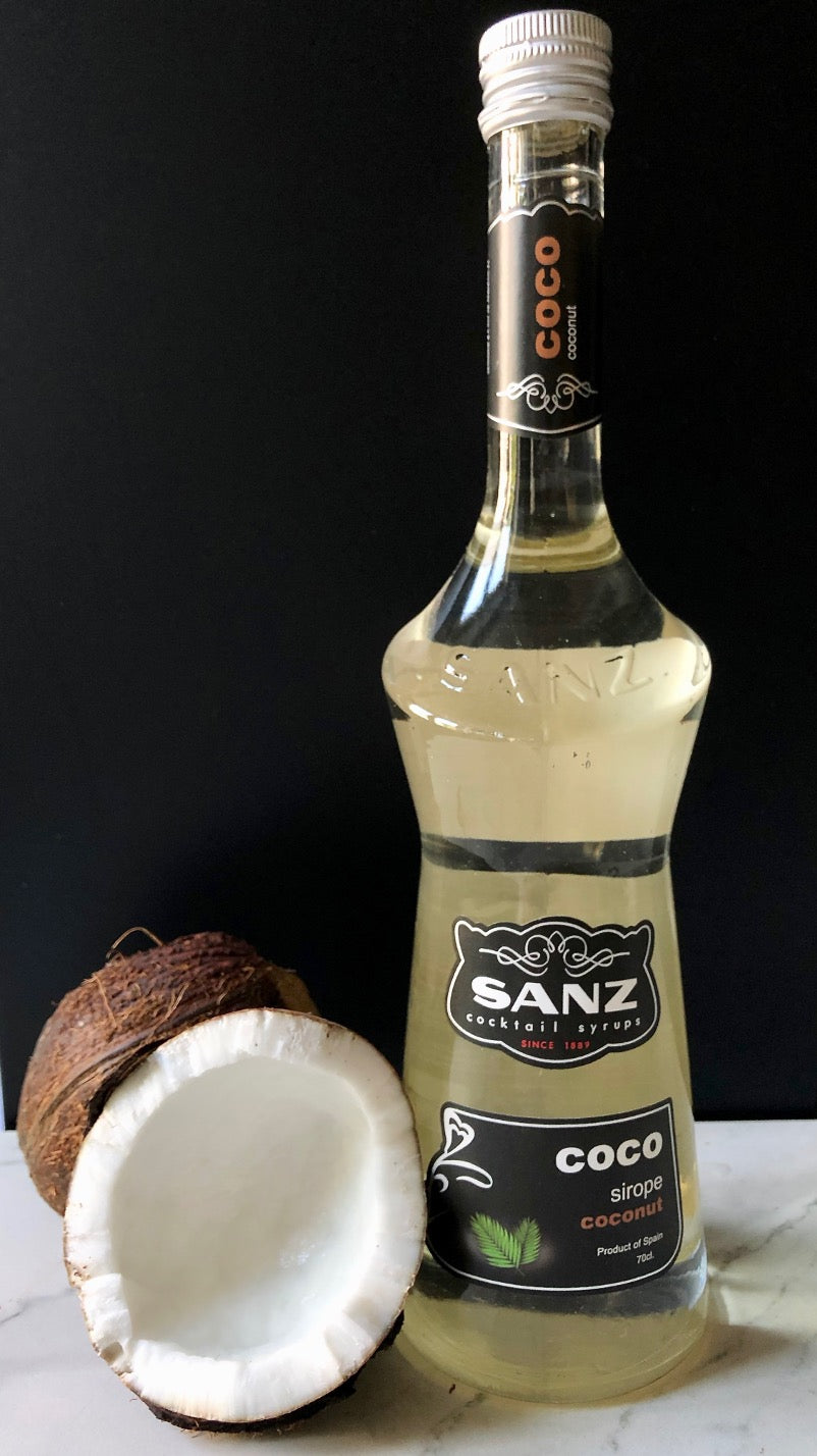 Flasche Kokossirup Sanz mit schwarzem Etikett, daneben eine halbierte Kokosnuss, dunkler Hintergrund, 70 cl