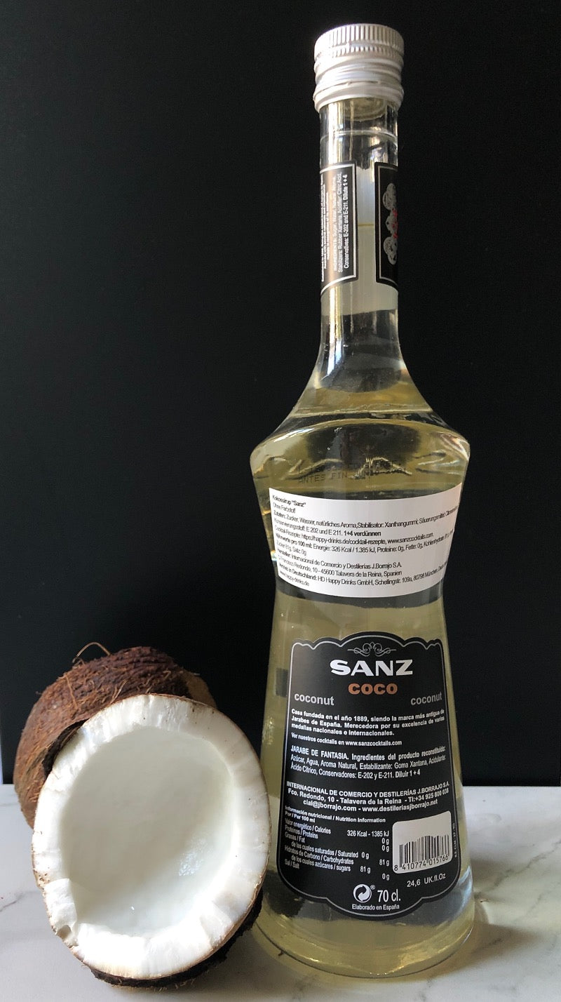 Rückseite der Kokossirup-Sanz-Flasche mit Zutatenliste und Barcode, daneben eine halbierte Kokosnuss, dunkler Hintergrund.