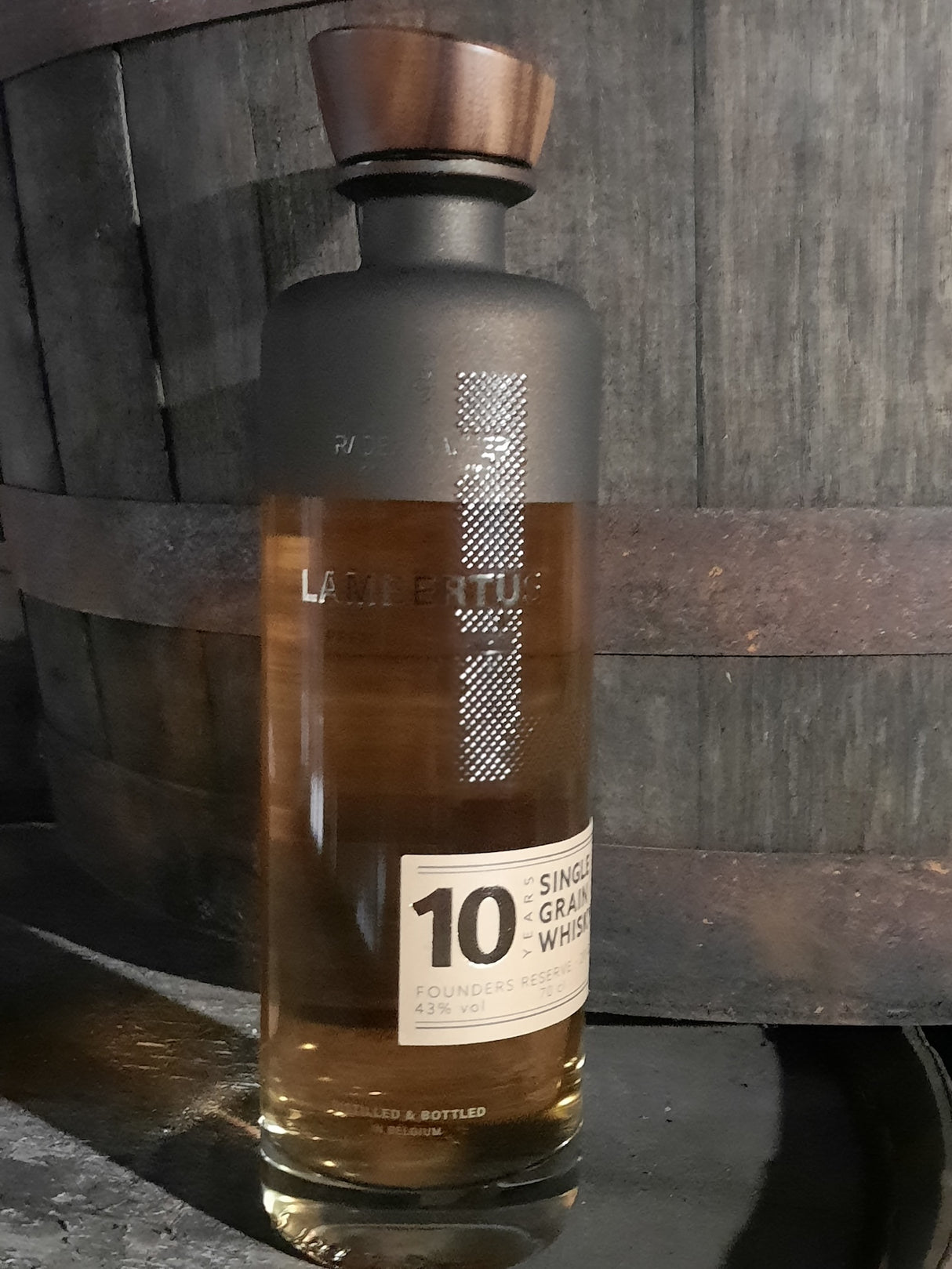 Lambertus 10 Years Founders Edition Whiskyflasche vor einem Holzfass, belgischer Single Grain Whisky mit Holzverschluss.