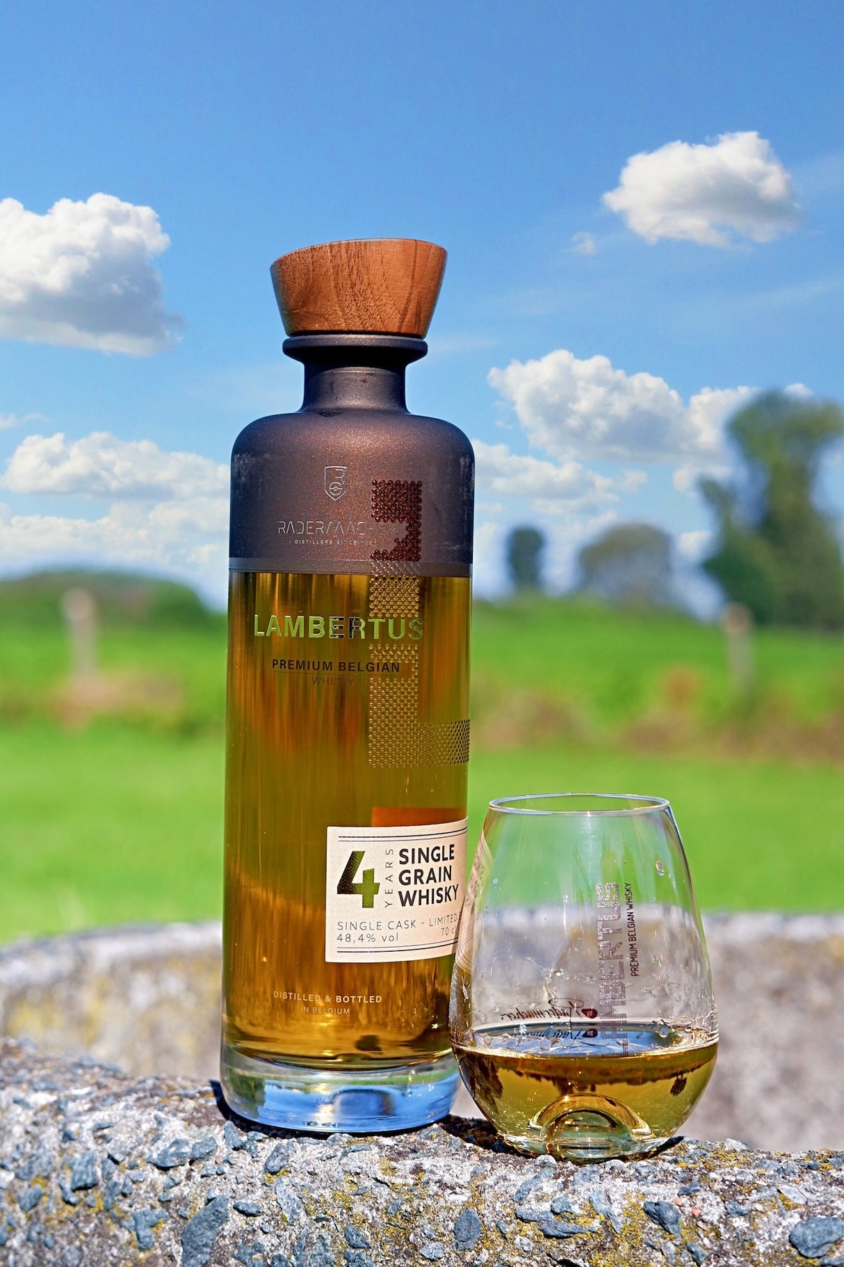 Lambertus Whiskyflasche und Glas auf Steinmauer vor blauem Himmel und grüner Landschaft