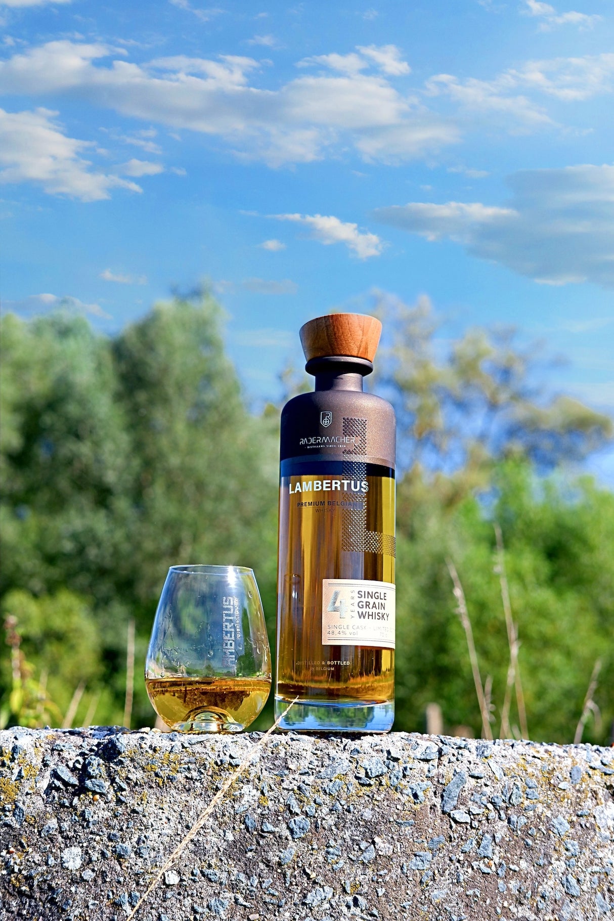 Lambertus Whiskyflasche und gefülltes Glas auf einer Steinmauer vor blauem Himmel und grünen Bäumen