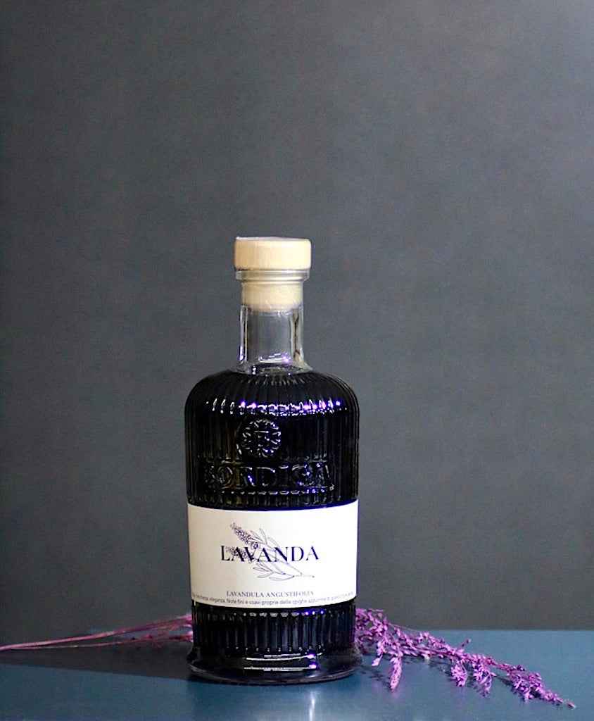 Flasche Bordiga Lavanda Likör mit cremefarbenem Etikett und Lavendelzweig auf glänzendem Tisch vor grauem Hintergrund.