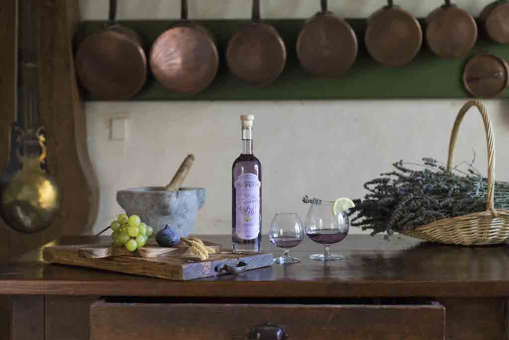 Flasche Fleur de Lavande Likör in rustikaler Küche mit Gläsern, Trauben, Feige, Korb Lavendel und Kupferpfannen im Hintergrund