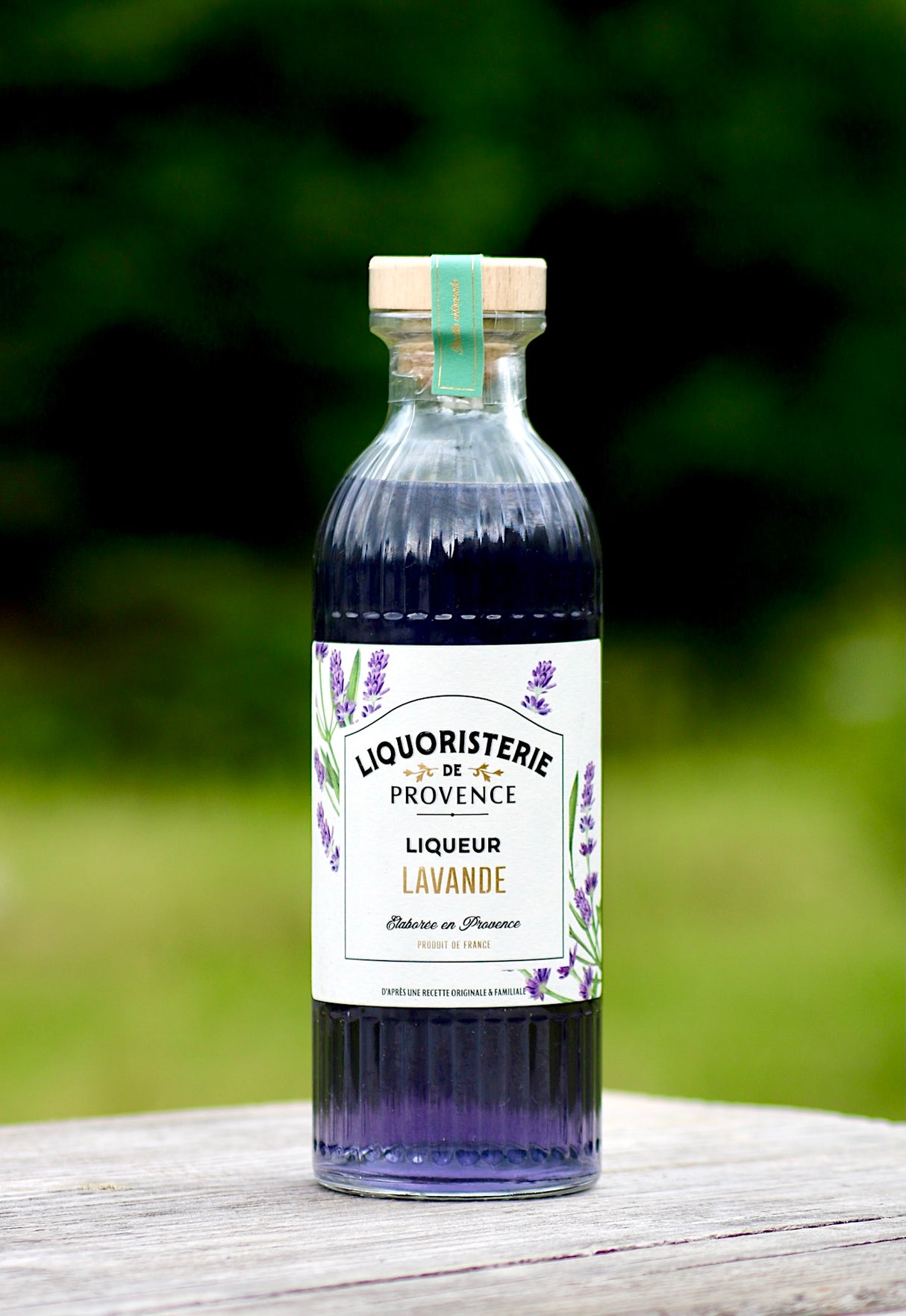 Lavendellikör Liquoristerie de Provence Flasche auf hellem Holztisch vor grünem Hintergrund