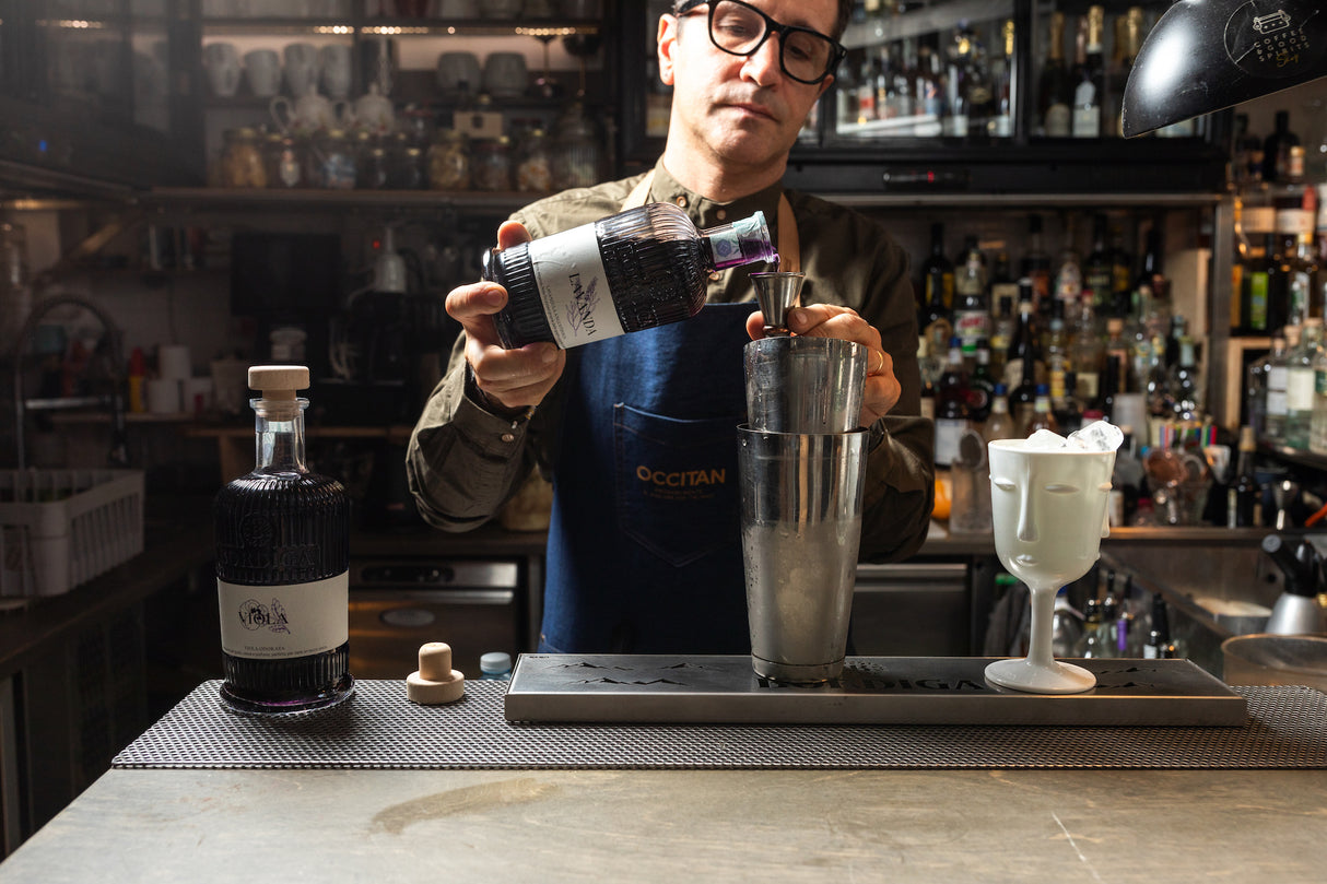 Bartender in einer Bar gießt Lavendellikör Bordiga aus einer Flasche in einen Messbecher, Cocktailzubereitung mit Metallshaker und Glas.