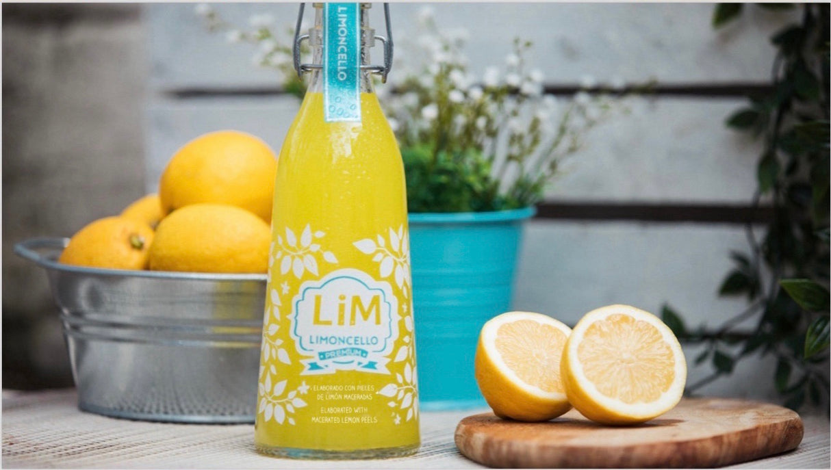 Flasche Lim Limoncello mit frischen Zitronen auf Holzbrett, im Hintergrund Metallschale und Blumentopf.
