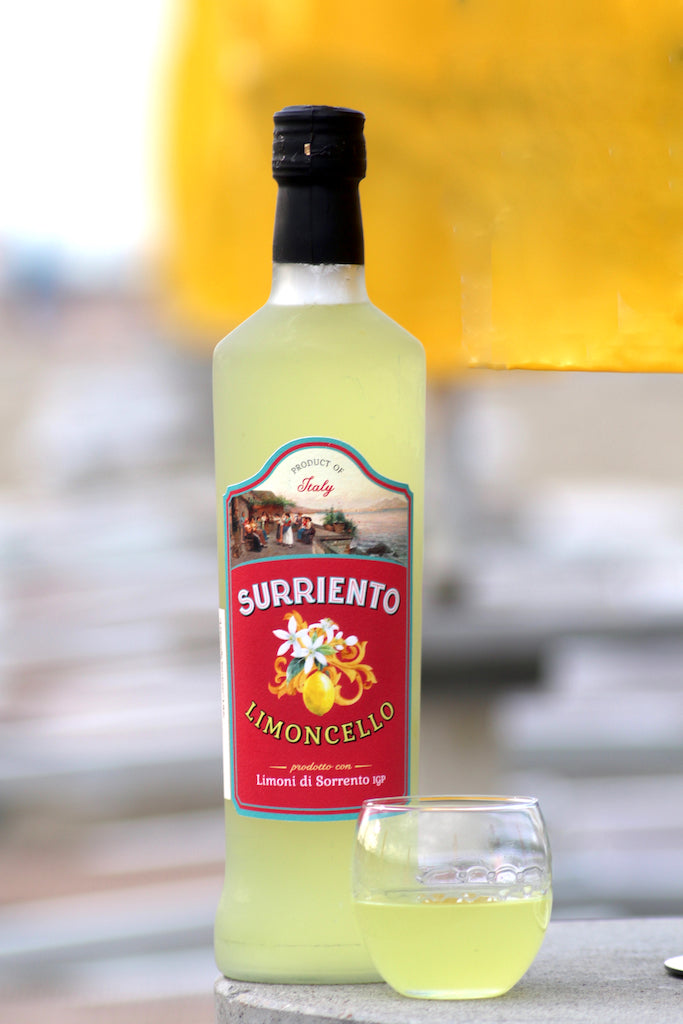Flasche Surriento Limoncello mit rotem Etikett und Glas mit gelbem Likör davor, vor unscharfem Hintergrund mit gelbem Element.