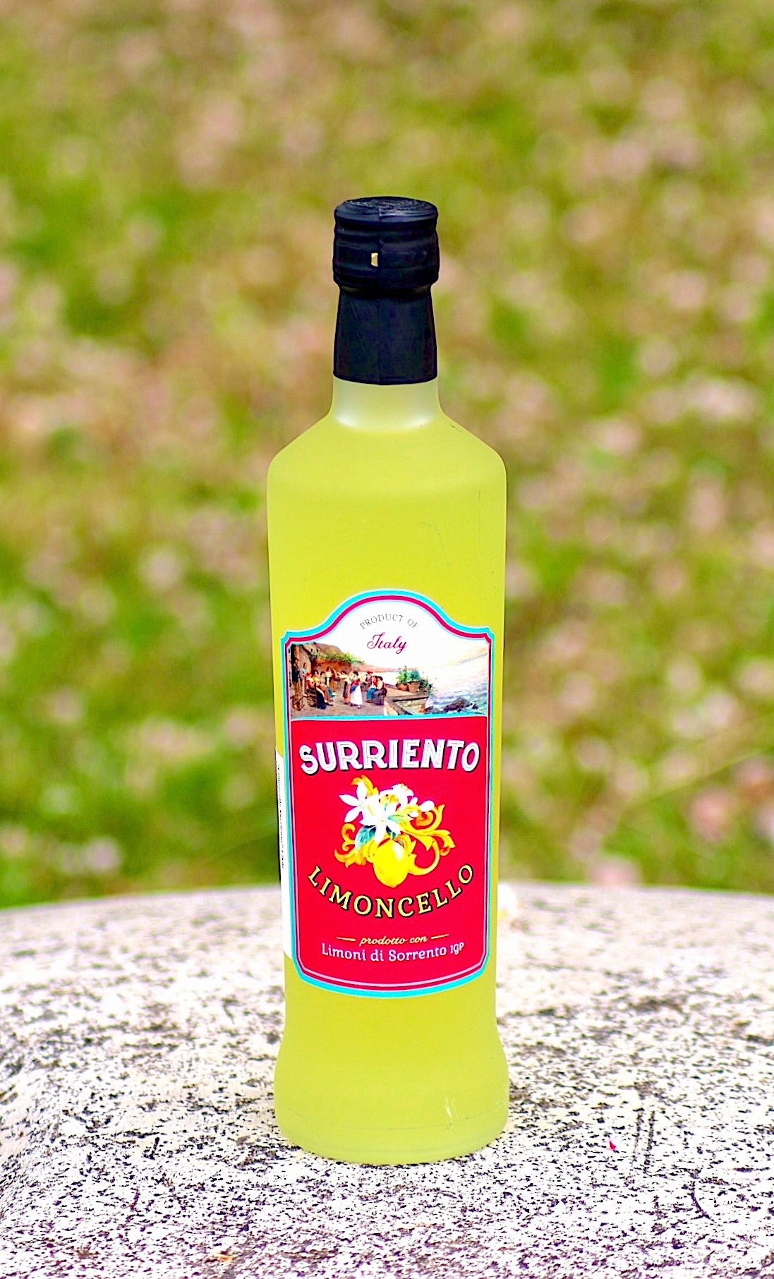 Flasche Surriento Limoncello mit rotem Etikett auf heller, strukturierter Oberfläche, verschwommener grüner Hintergrund mit rosa Akzenten.