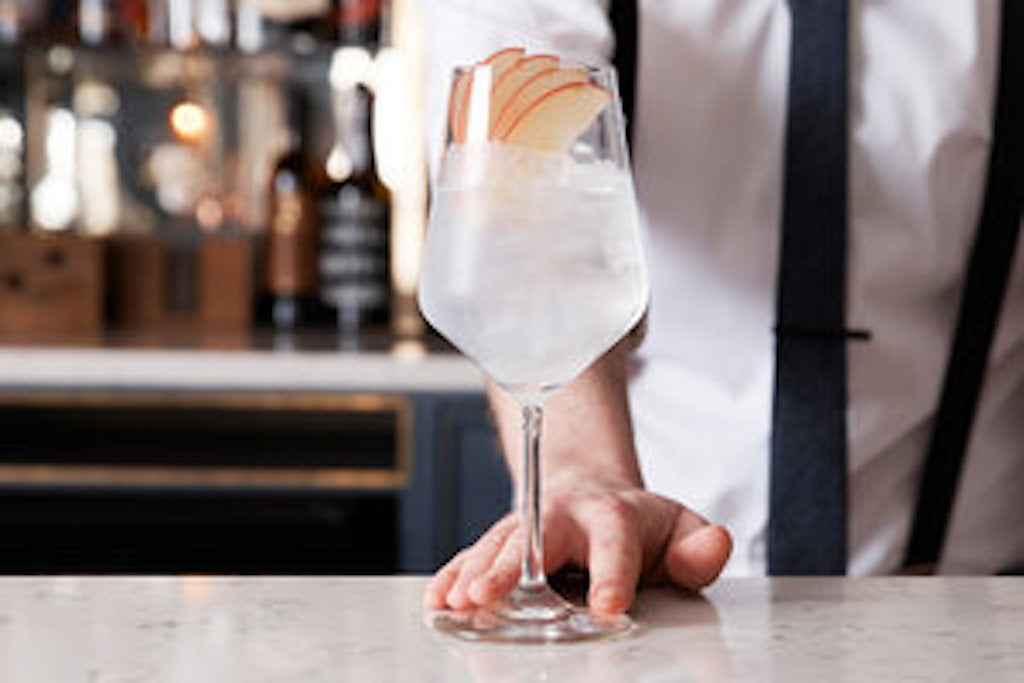 Elegantes Cocktailglas mit einer Gin-Tonic-Kreation, dekoriert mit frischen Apfelscheiben, präsentiert von einem Barkeeper im Hintergrund.