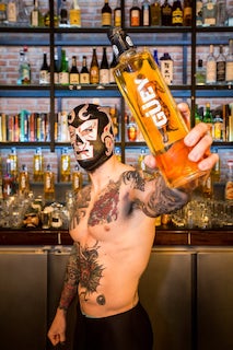 Lucha Libre-Kämpfer mit Maske präsentiert eine Flasche Güey Hot Spicy Tequila vor einer gut bestückten Bar.