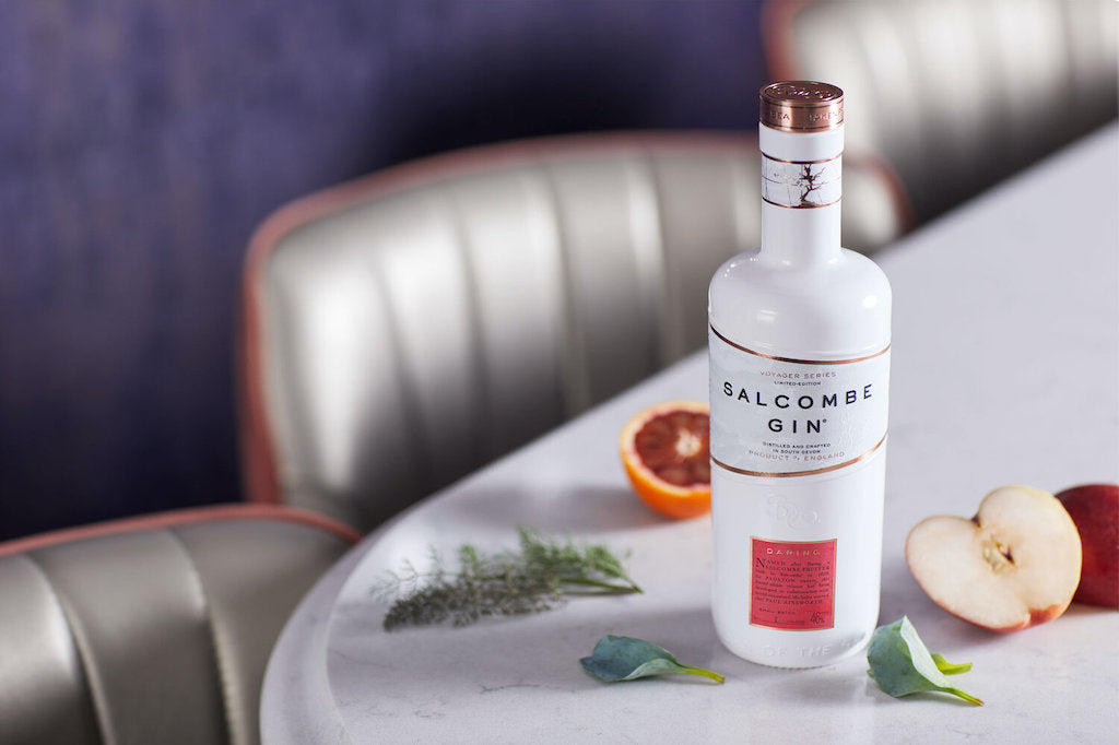 Salcombe Gin Daring, weiße Flasche mit rotem Etikett, präsentiert auf einem Marmortisch mit Apfelstücken, Kräutern und Blutorange.