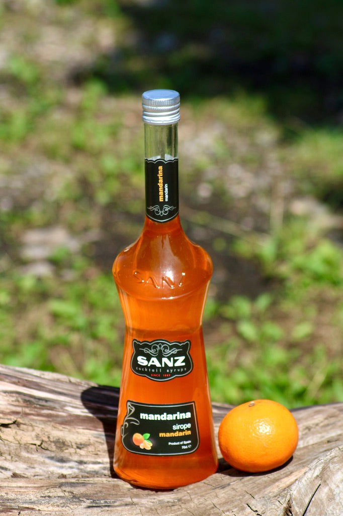 Flasche Mandarinen-Sirup von Sanz in orangener Farbe, mit schwarzem Etikett und silbernem Verschluss auf einem Holzstamm neben einer Mandarine.