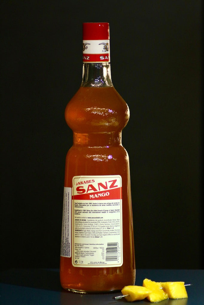 Mango Sirup Sanz 1L Flasche, Rückseite mit Etikett und Zutatenliste, daneben frische Mangostücke auf einem Spieß.