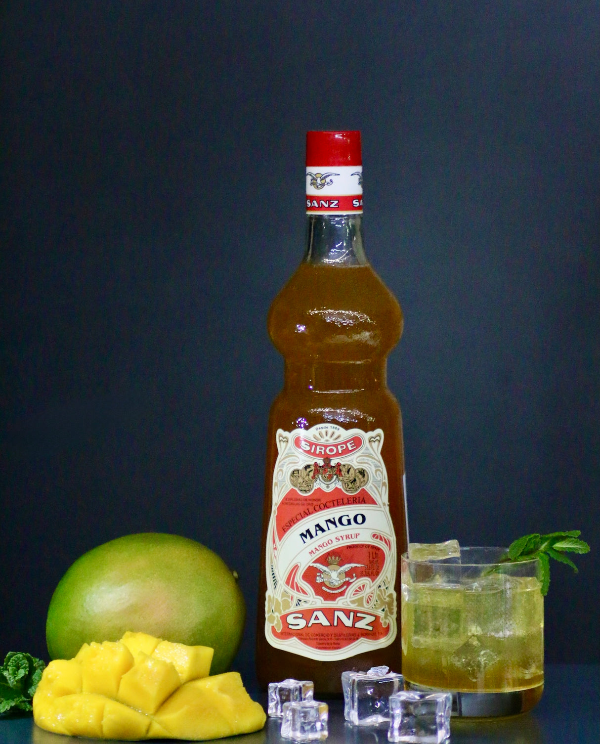 Mangosirup Sanz 1L mit frischer Mango, Eiswürfeln und Cocktail auf dunklem Hintergrund.