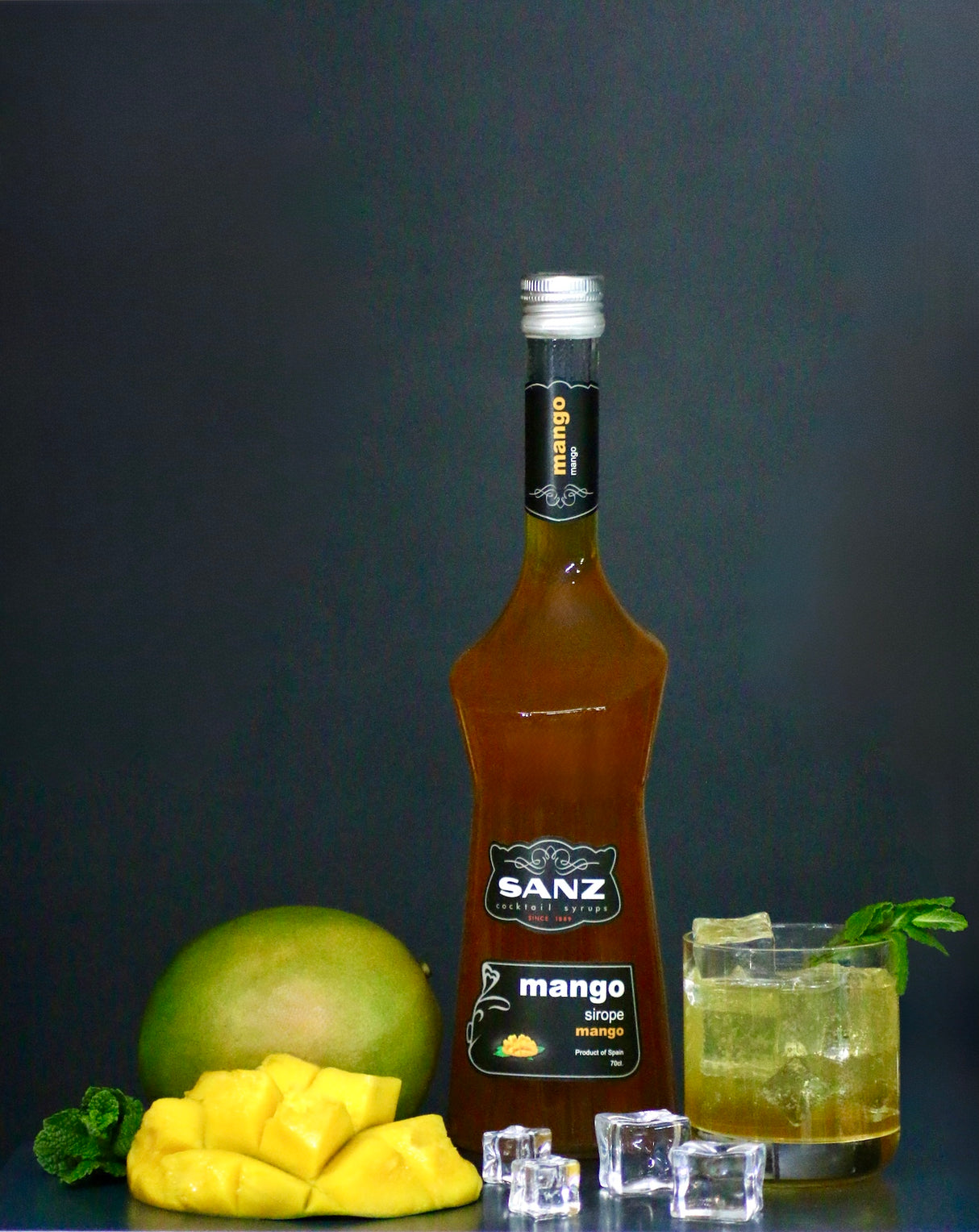 Mangosirup Sanz 0,7L mit frischer Mango, Eiswürfeln und Mango Tonic Cocktail auf dunklem Hintergrund.