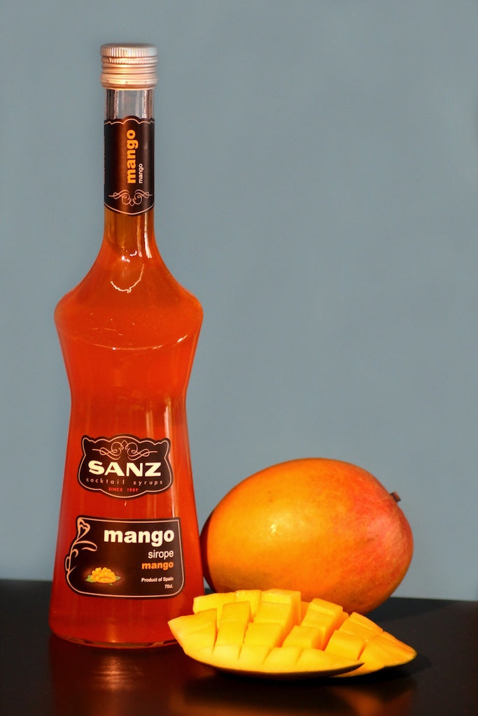 Mangosirup Sanz in einer 0,7-Liter-Flasche, daneben eine ganze Mango und ein aufgeschnittenes Mangostück vor blauem Hintergrund.