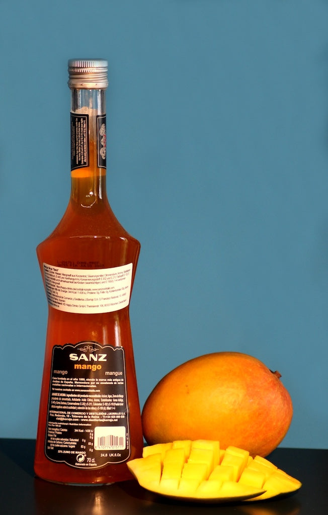 Mangosirup Sanz in einer 0,7-Liter-Flasche, Rückseite mit Etikett sichtbar, daneben eine ganze Mango und ein aufgeschnittenes Mangostück.