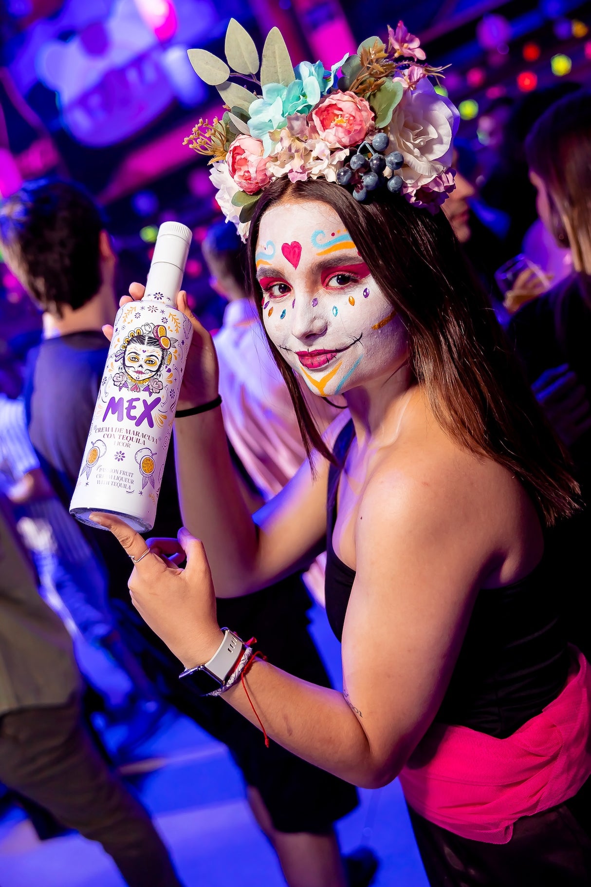 Frau mit farbenfrohem Gesichtsmuster und Blumenkranz hält eine Flasche Mex Maracuja-Tequila Sahnelikör auf einer Party.