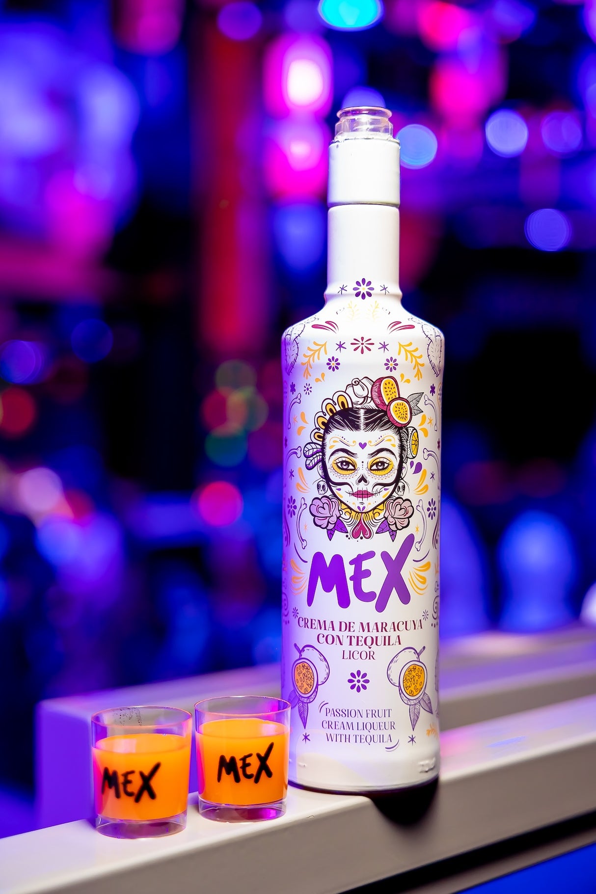Flasche Mex Maracuja-Tequila Sahnelikör mit zwei gefüllten Shotgläsern vor unscharfem, farbig beleuchtetem Hintergrund.