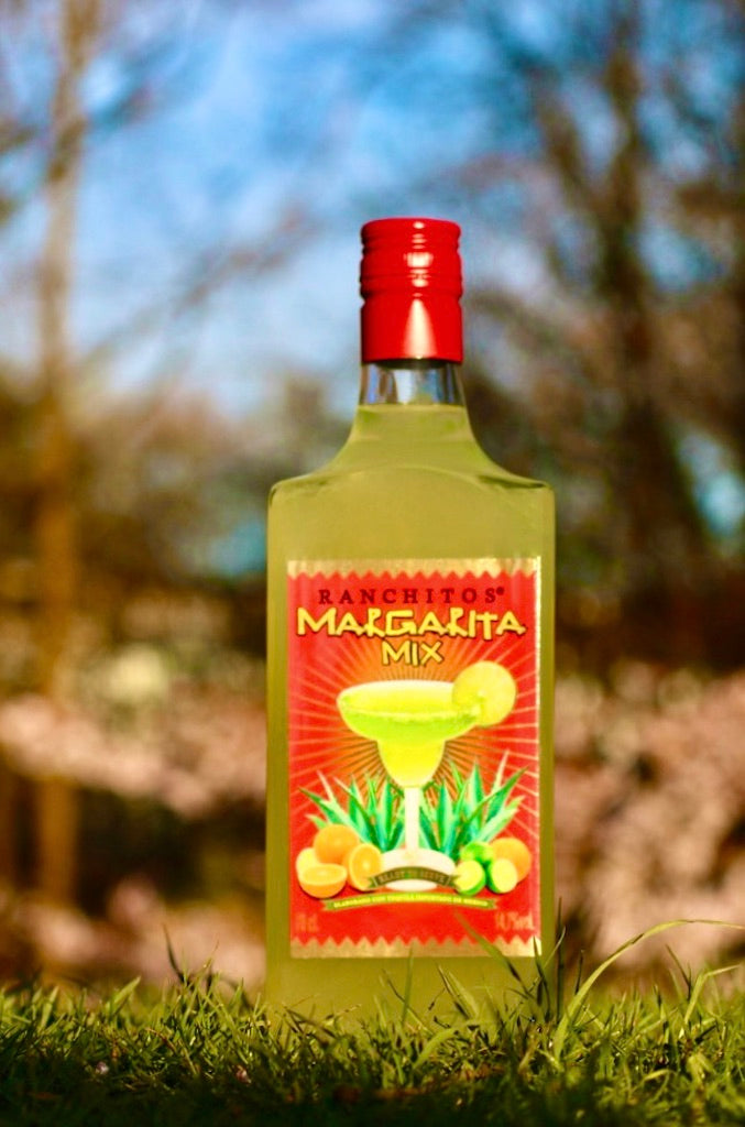 Flasche Ranchitos Margarita Mix, stehend auf Gras mit leuchtend rotem Etikett, das eine Margarita zeigt, vor einem natürlichen Hintergrund.