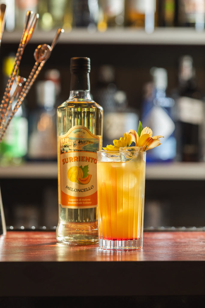 Flasche Surriento Meloncello neben einem Meloncello Sour Cocktail im Glas mit Eiswürfeln und gelber Blütendekoration auf einer Bar.