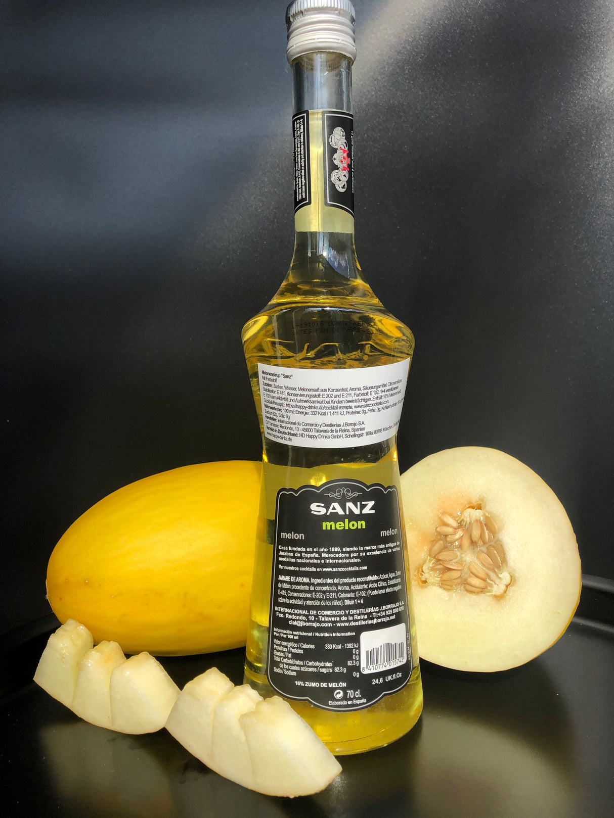Rückseite der Sanz Melonensirup-Flasche mit Produktinformationen, daneben Honigmelone und Melonenstücke vor schwarzem Hintergrund.