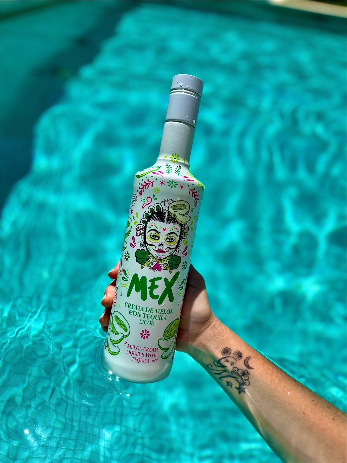Hand mit Tattoo hält eine Flasche Mex Melonen-Tequila Sahnelikör über türkisblauem Poolwasser.