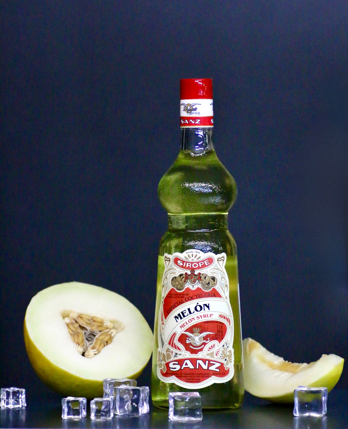 Melonensirup Sanz 1L, gelber Sirup in Flasche mit rotem Etikett, daneben Melonenscheiben und Eiswürfel vor schwarzem Hintergrund.