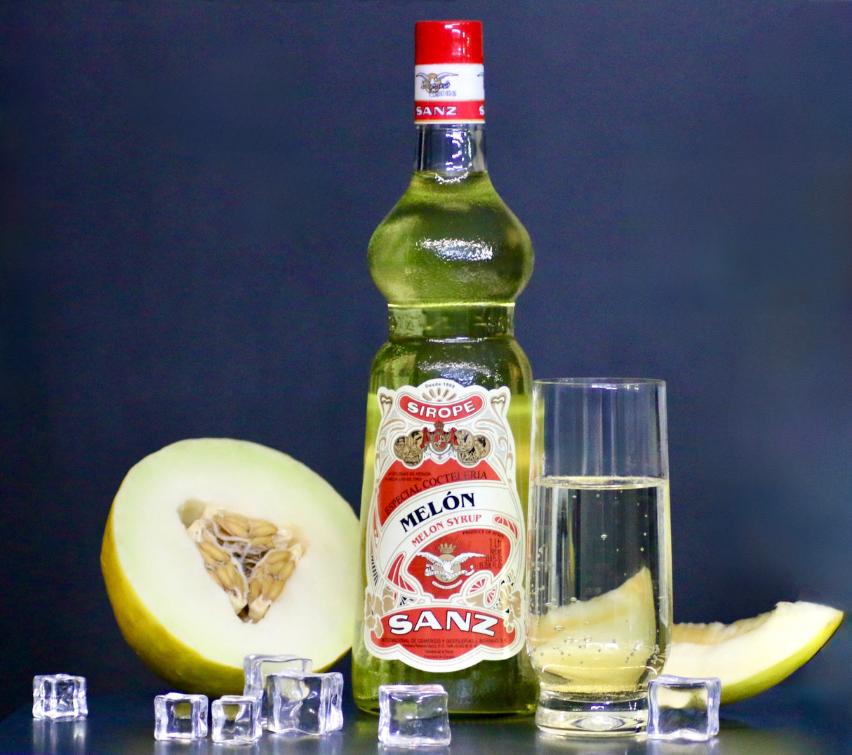 Melonensirup Sanz 1L, gelber Sirup in Flasche mit rotem Etikett, Glas mit Sirup, Melonenscheiben und Eiswürfeln.
