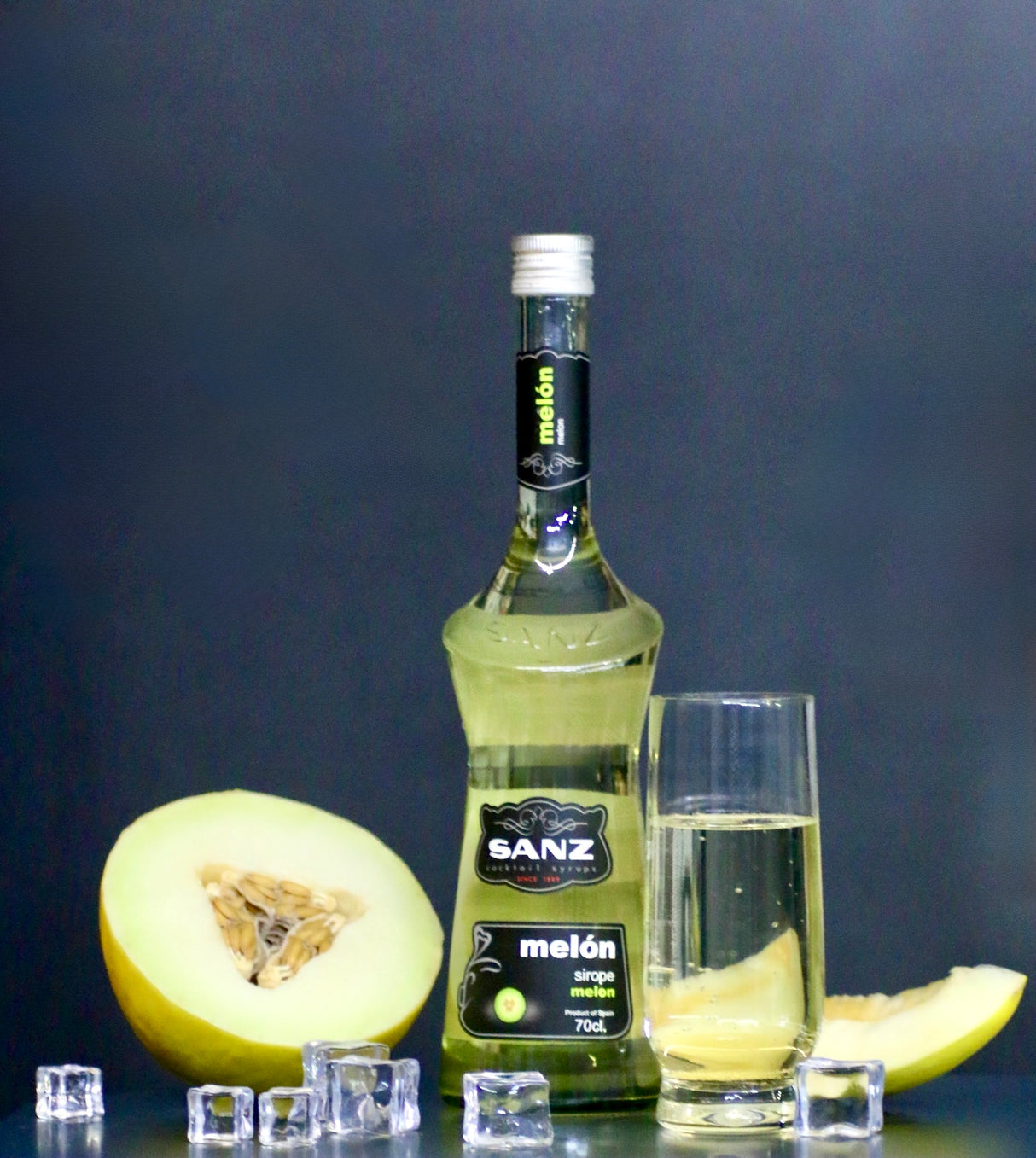 Melonensirup Sanz 700 ml, daneben ein Glas mit Sirup, Eiswürfel und gelbe Melonenscheiben vor schwarzem Hintergrund.