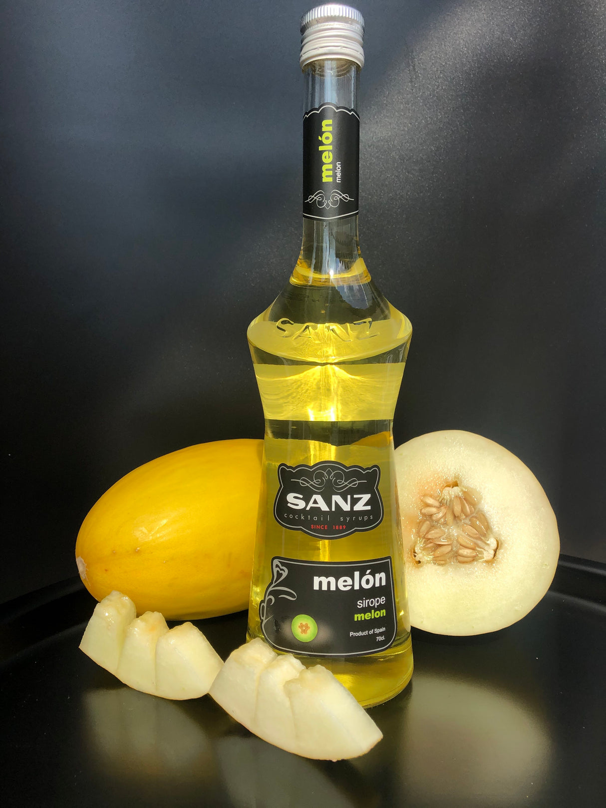 Flasche Sanz Melonensirup mit hellgelbem Inhalt, umgeben von einer Honigmelone und Melonenstücken vor schwarzem Hintergrund.