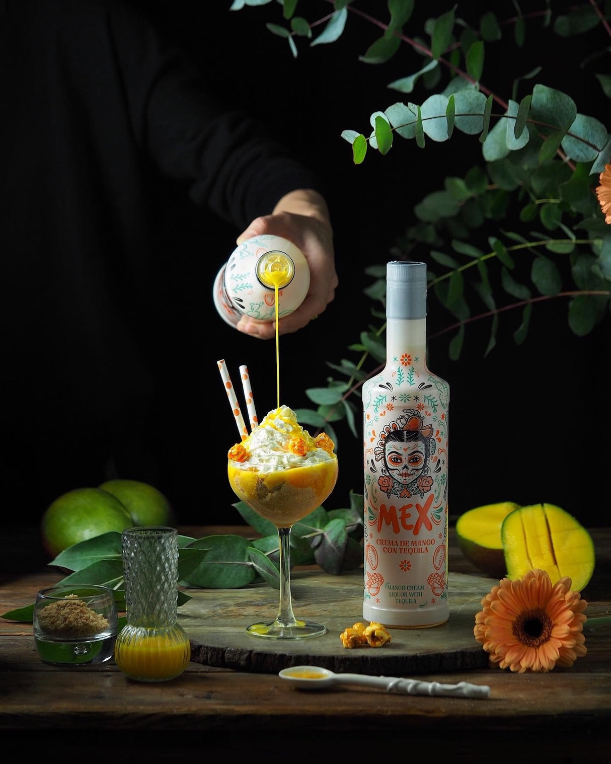 Mex Mango-Tequila Likör mit Sahne, kunstvoll serviert mit Dessert, frischen Mangos und dekorativen Elementen auf rustikalem Holztisch.