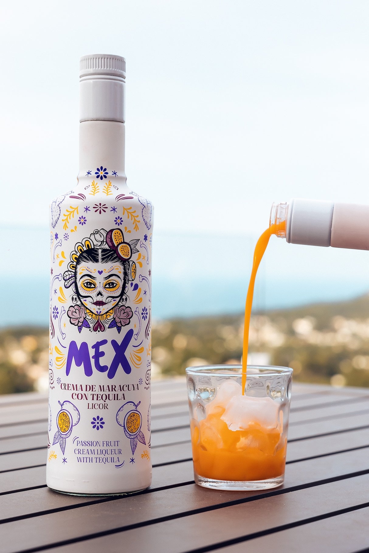Flasche Mex Maracuja-Tequila Sahnelikör auf Holztisch mit Meerblick, daneben Glas mit Eis, in das Likör aus einer weiteren Flasche fließt.