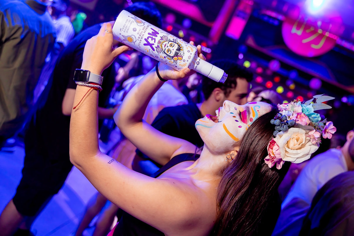 Frau mit kunstvoll bemaltem Gesicht und Blumenkranz hält eine Flasche Mex Maracuja-Tequila Sahnelikör auf einer Party.