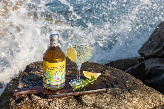 Nerses Yellow Gin & Tonic auf Fels mit Zitronenscheibe und Rosmarin, Flasche und Drink vor spritzender Meereskulisse.