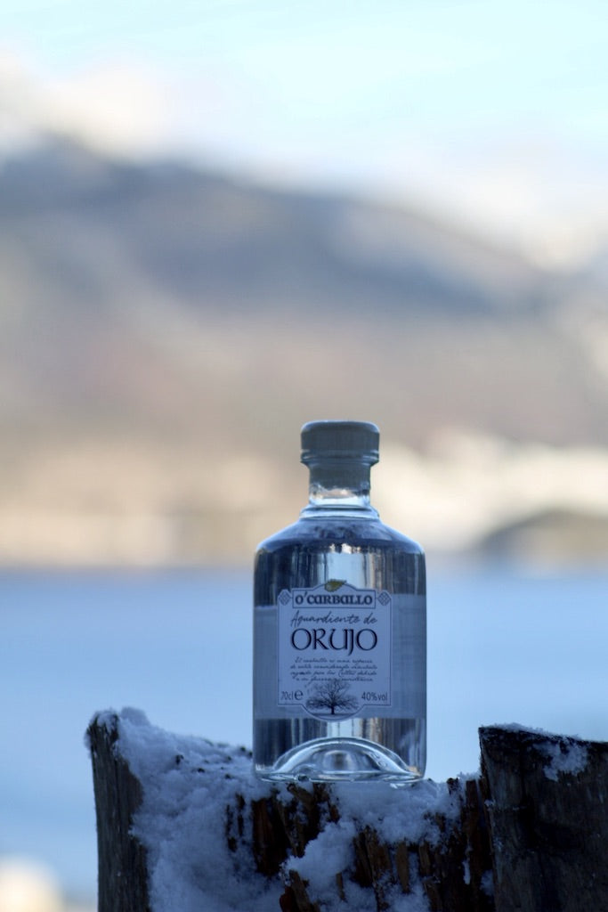 O’Carballo Orujo Flasche im Schnee auf Holzstumpf mit Berg- und Seepanorama im Hintergrund, klarer Tresterschnaps, 70 cl, 40 % vol.