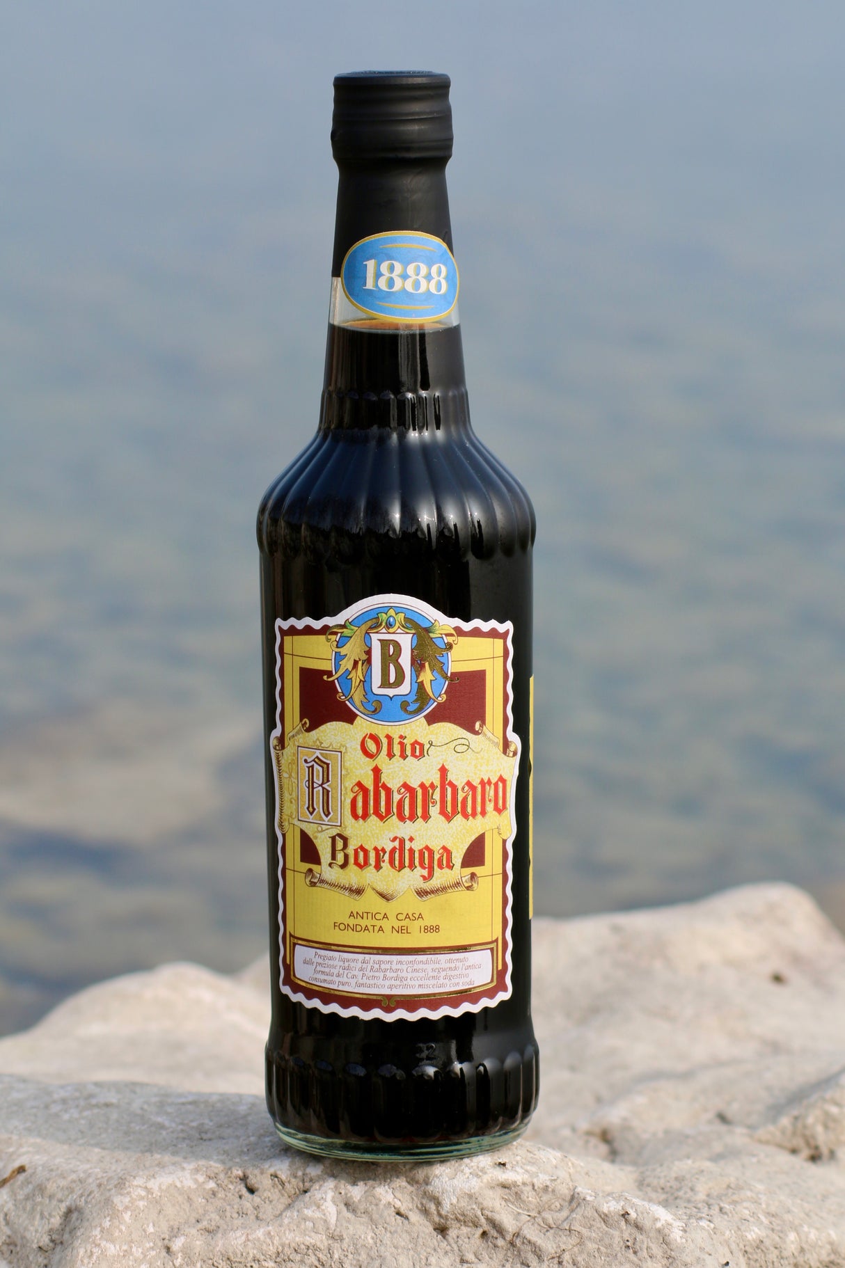 Flasche Olio di Rabarbaro Bordiga Rhabarber-Likör auf Felsen, dekoratives Etikett im Vordergrund, Wasser im Hintergrund.