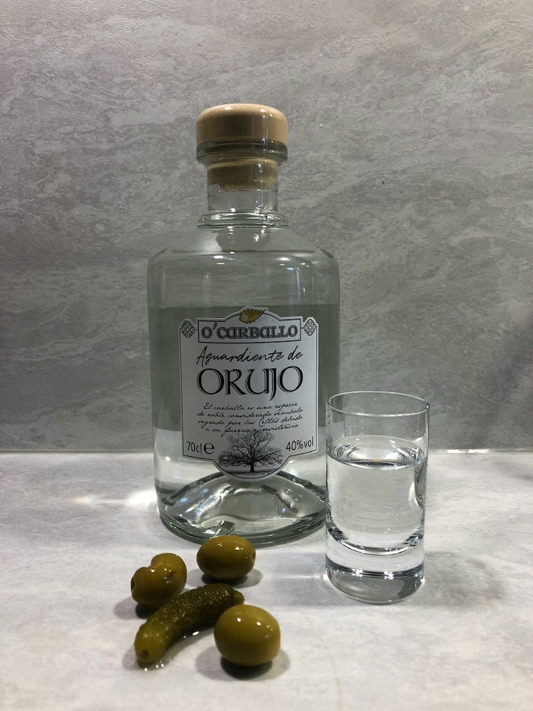 O’Carballo Orujo Flasche mit gefülltem Schnapsglas, grünen Oliven und Cornichon auf hellem Untergrund, 70 cl, 40 % vol.