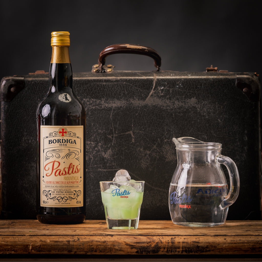 Bordiga Pastis-Flasche, Glas mit Pastis und Eiswürfeln, Wasserkaraffe, vor altem Koffer als Hintergrund