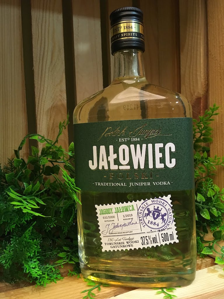 Jalowiec Polski Wacholder-Wodka in grüner Etikettflasche, stehend vor Holzrückwand mit grünen Pflanzen.
