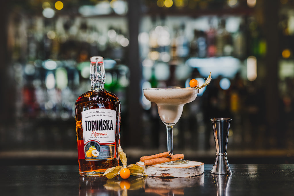 Flasche Toruńska Quitte Likör neben einem eleganten Cocktailglas mit garnierter Physalis und Zimtstange, vor einer unscharfen Bar-Hintergrundkulisse.