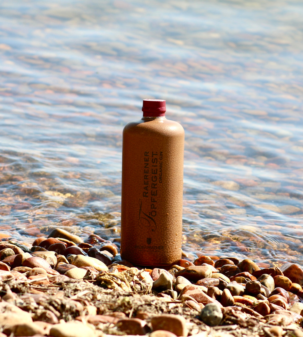 Tonflasche Raerener Töpfergeist Bio-Gin auf Kieselstrand vor klarem Wasser mit sanften Wellen im Hintergrund.