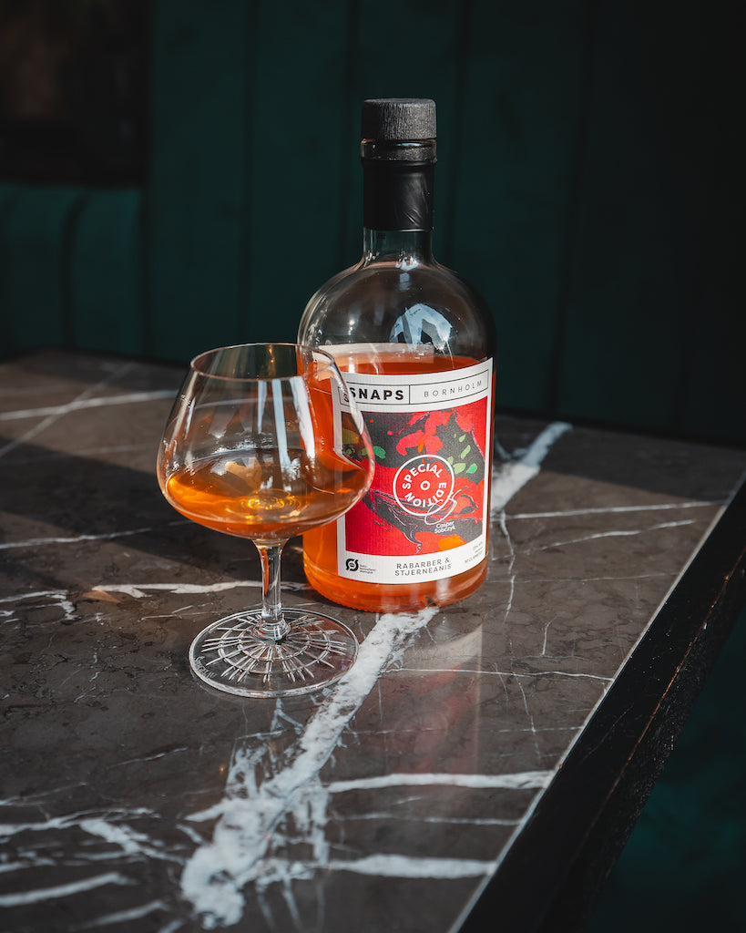 Bio-Schnaps Bornholm Rhabarber & Sternanis, Flasche auf Marmortisch mit gefülltem Glas, dunkler Hintergrund mit grüner Polsterung.