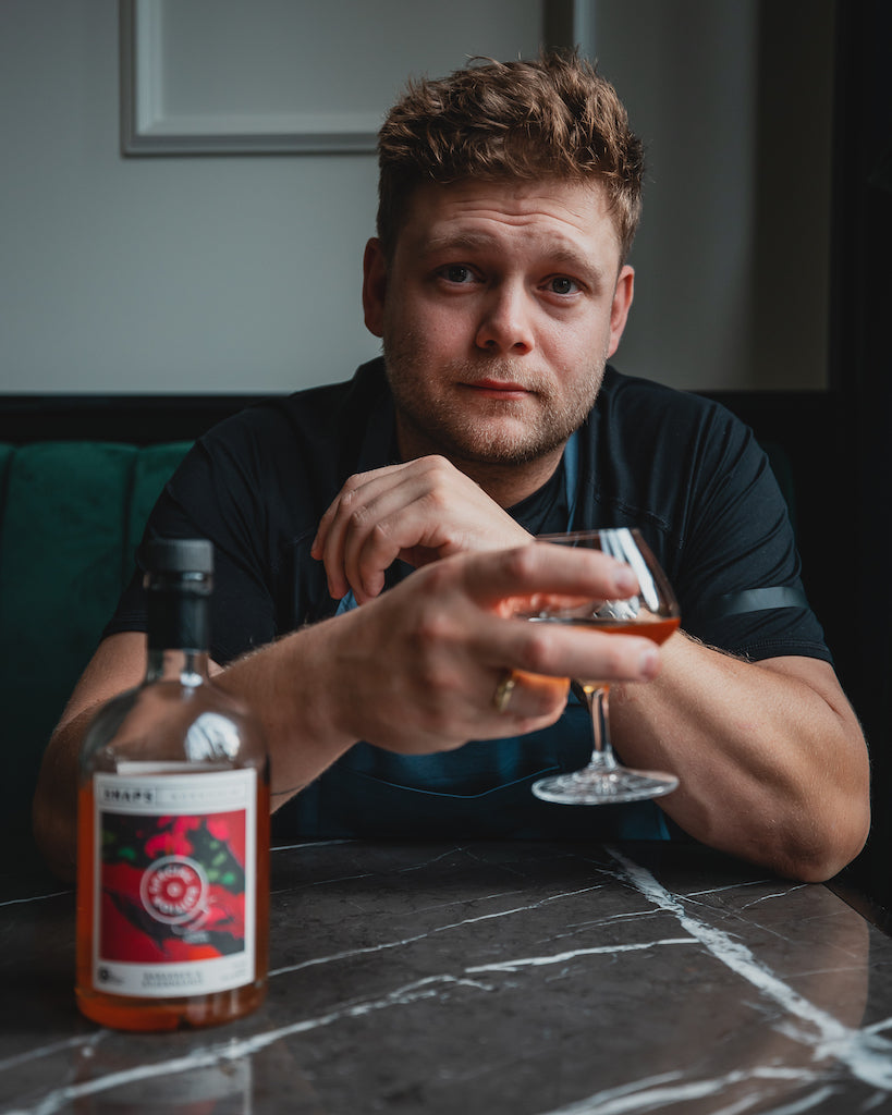 Koch mit blauer Schürze hält ein Glas Bio-Schnaps Bornholm Rhabarber & Sternanis, Flasche auf Marmortisch, gemütliche Bar-Atmosphäre.