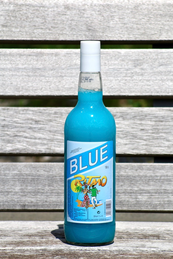 Flasche Ritmo Blue 1 L mit blauem Getränkekonzentrat und weißem Verschluss auf Holzbank vor grüner Wiese