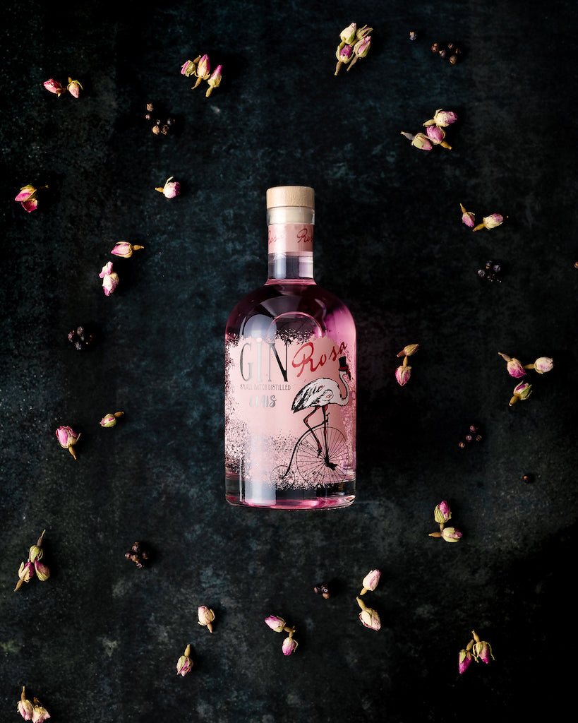 Flasche Ciais Gin Rosa von Bordiga auf dunklem Untergrund, umgeben von rosa Rosenknospen und Wacholderbeeren.