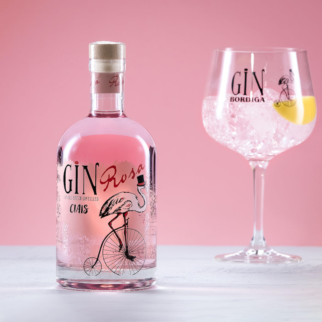 Flasche Ciais Gin Rosa von Bordiga vor rosa Hintergrund, daneben ein Ballonglas mit Gin Tonic, Eiswürfeln und Zitronenzeste.