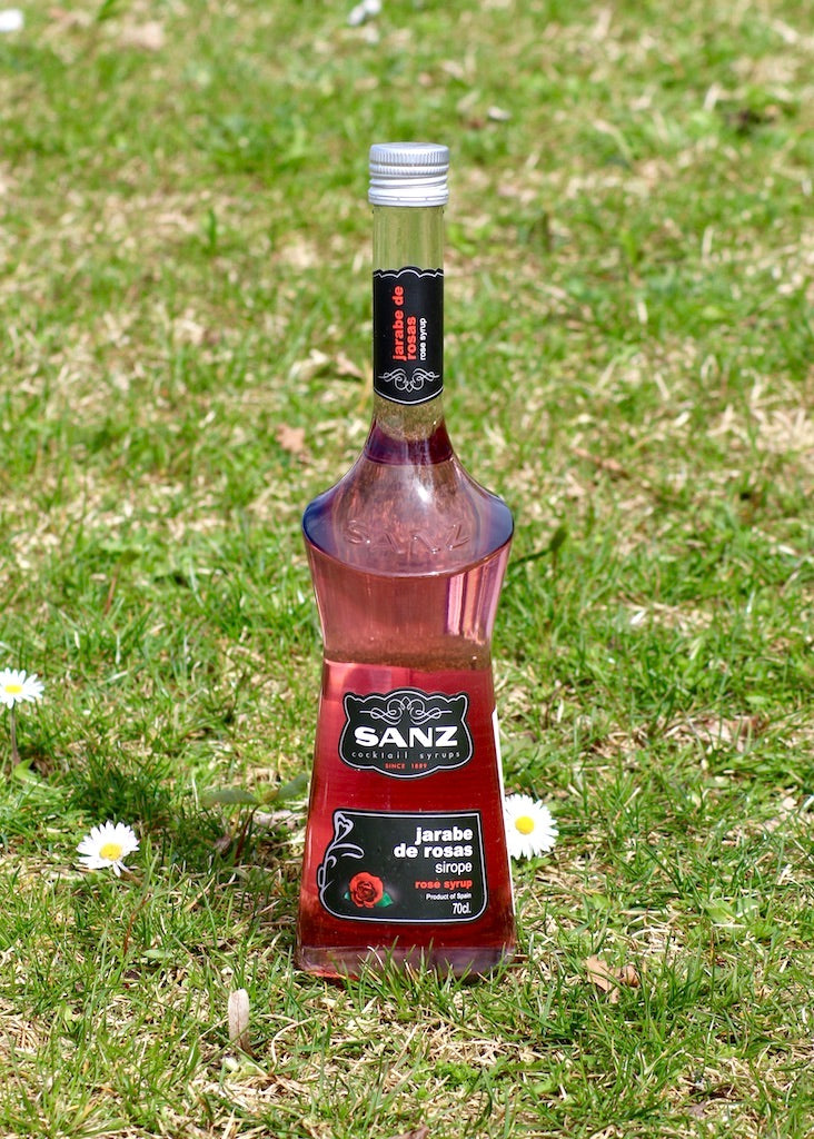 Sanz Rosensirup 700 ml, elegante Flasche mit schwarzem Etikett, auf einer grünen Wiese mit Gänseblümchen platziert.