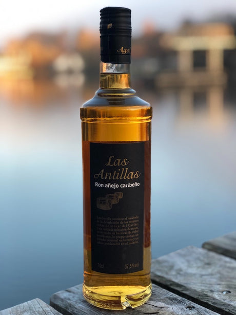 Flasche Las Antillas Ron Añejo Caribeño auf Holzfläche vor ruhigem See und unscharfem Hintergrund, 70 cl, 37,5 % Vol. Alkohol.