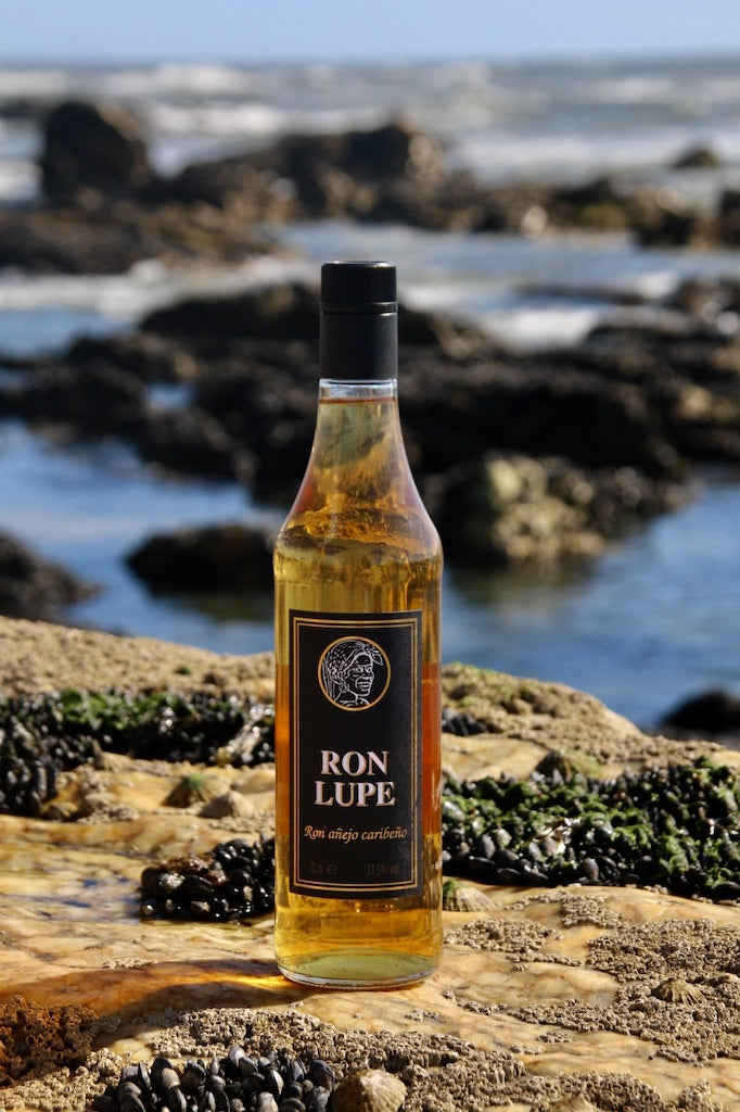Rum Añejo Lupe mit schwarzem Etikett, goldbraune Flasche auf Felsen am Meer mit Wellen im Hintergrund.